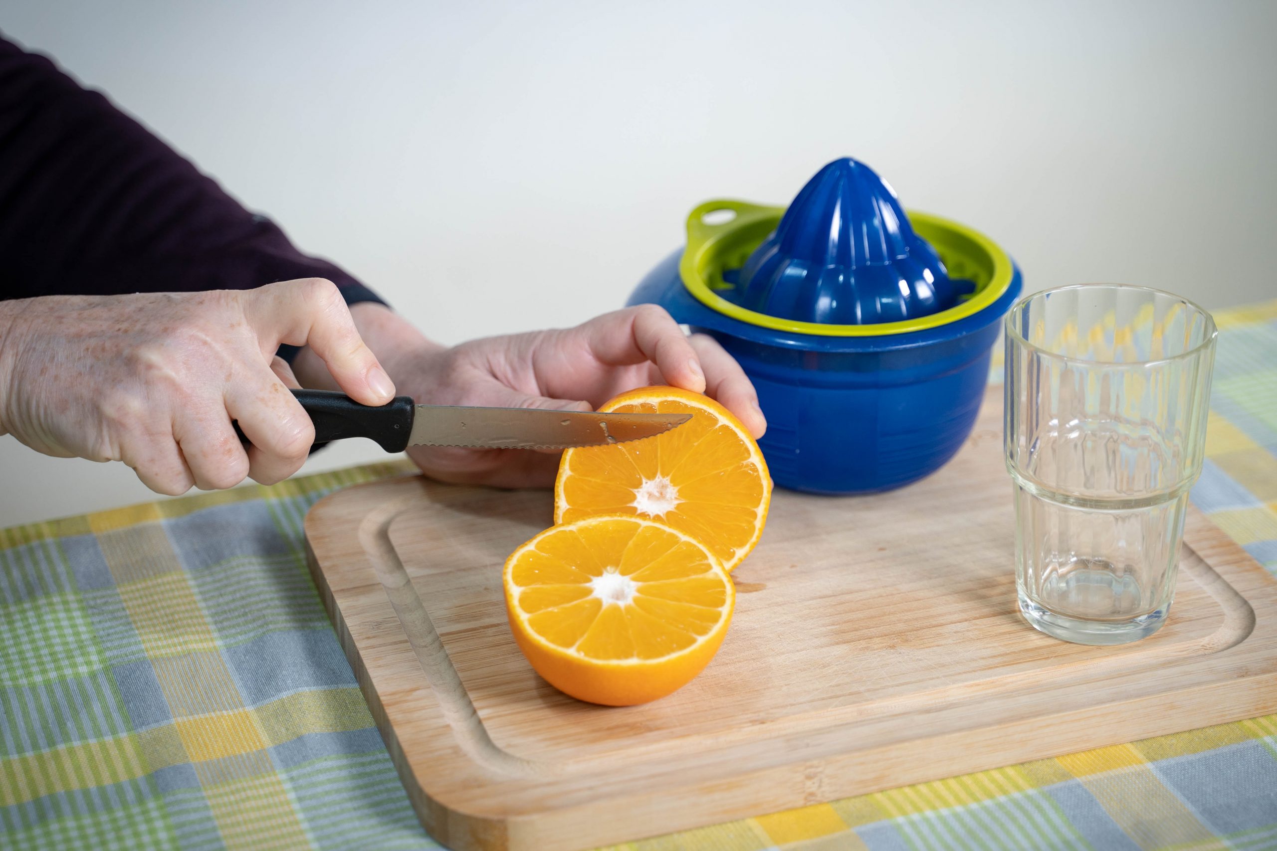 .ידיים חוצות תפוז לשני חצאים באמצעות סכין, על גבי קרש חיתוך שנמצא על שולחן. על גבי הקרש, מסחטת מיץ וכוס זכוכית ריקה