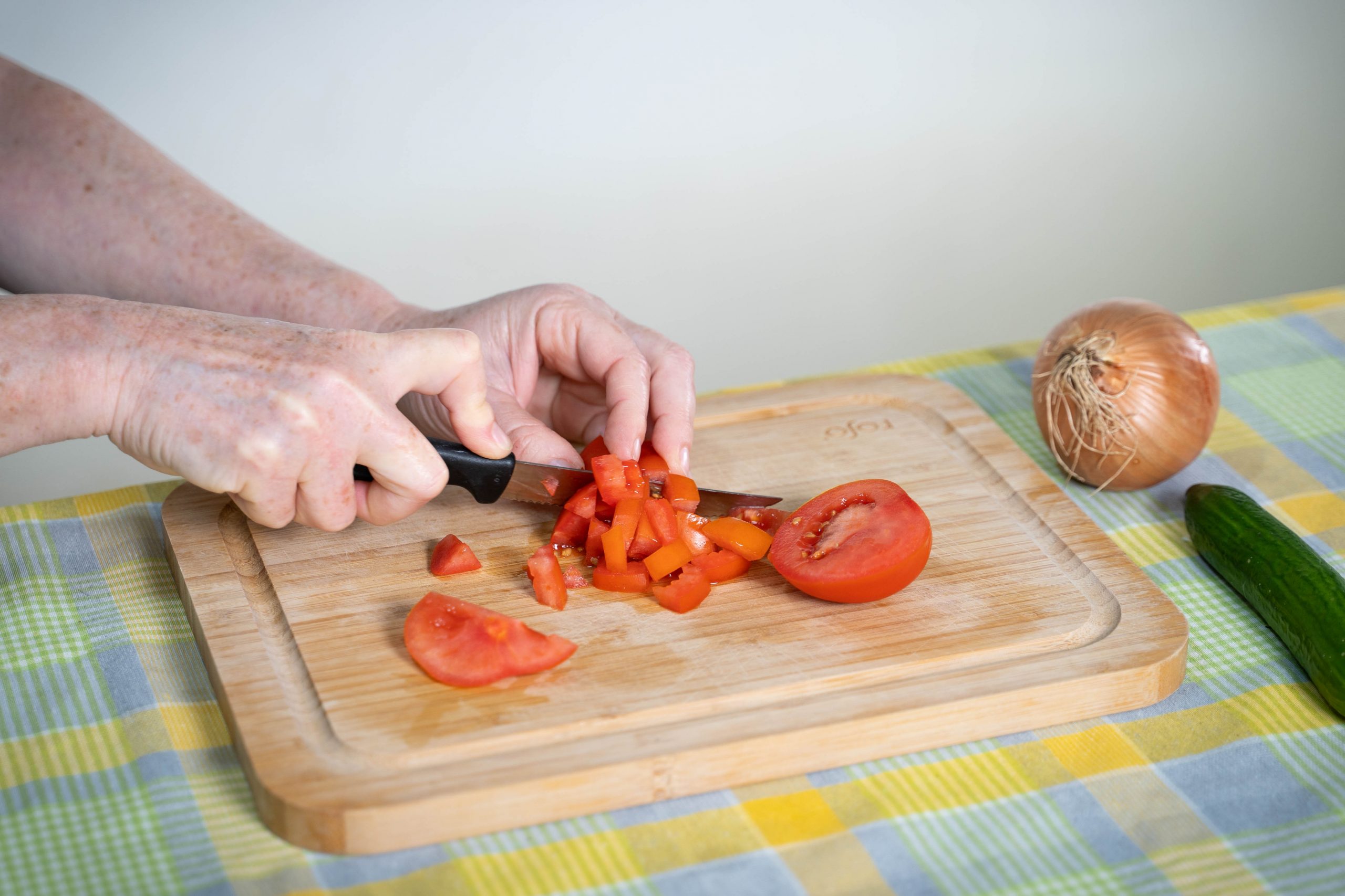 .ידיים חותכות עגבניה לסלט באמצעות סכין, על גבי קרש חיתוך. לצד הקרש, על השולחן, מונחים מלפפון ובצל