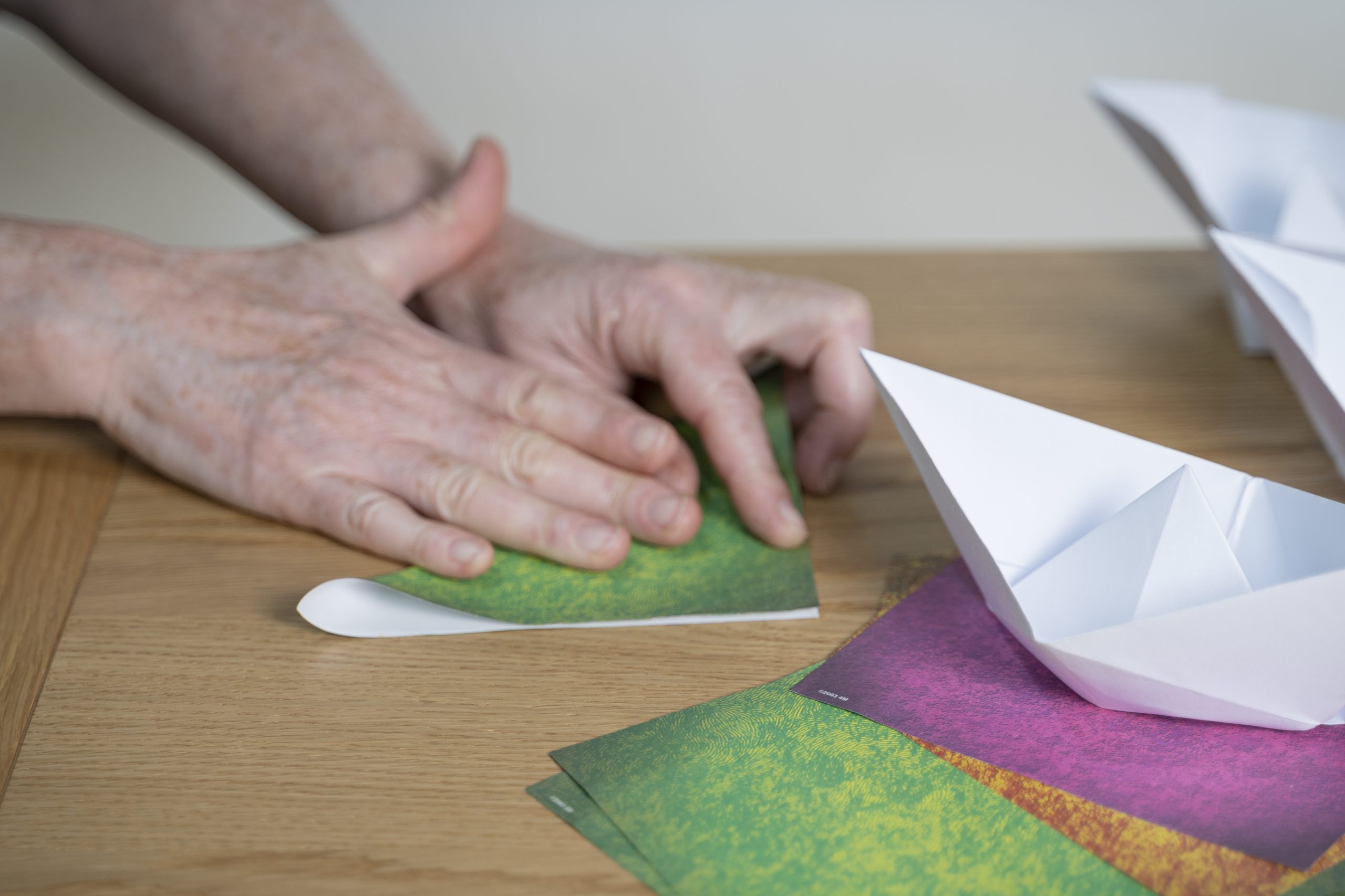 .זוג ידיים מקפלות נייר אוריגמי לצורת משולש. על השולחן ברקע ניירות אוריגמי צבעוניים ולבנים נוספים