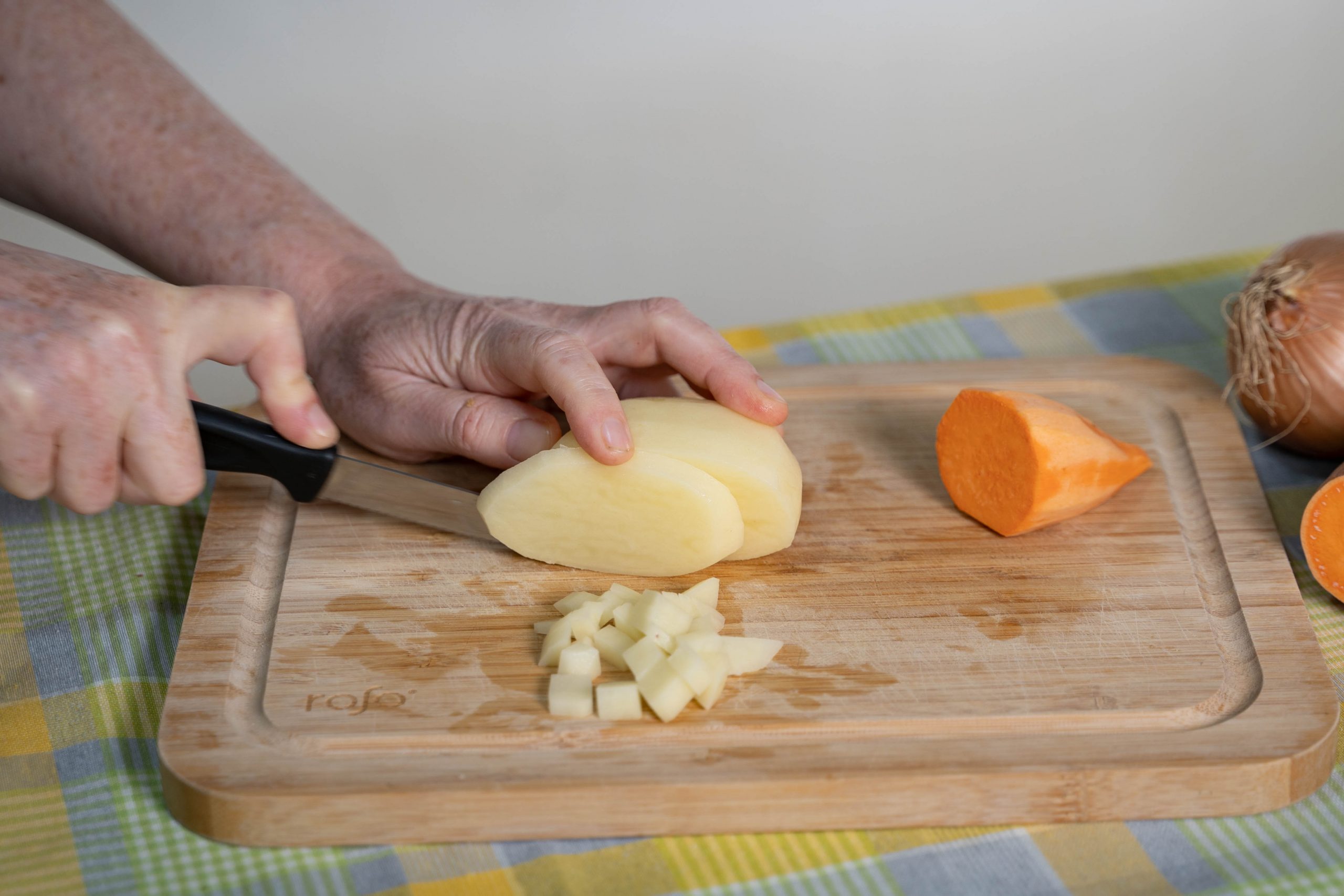 .יד חותכת תפוח אדמה לפרוסות באמצעות סכין, על גבי קרש חיתוך. לצד הפרוסות, קוביות תפוחי אדמה וחצי בטטה. ברקע, על השולחן, בצל