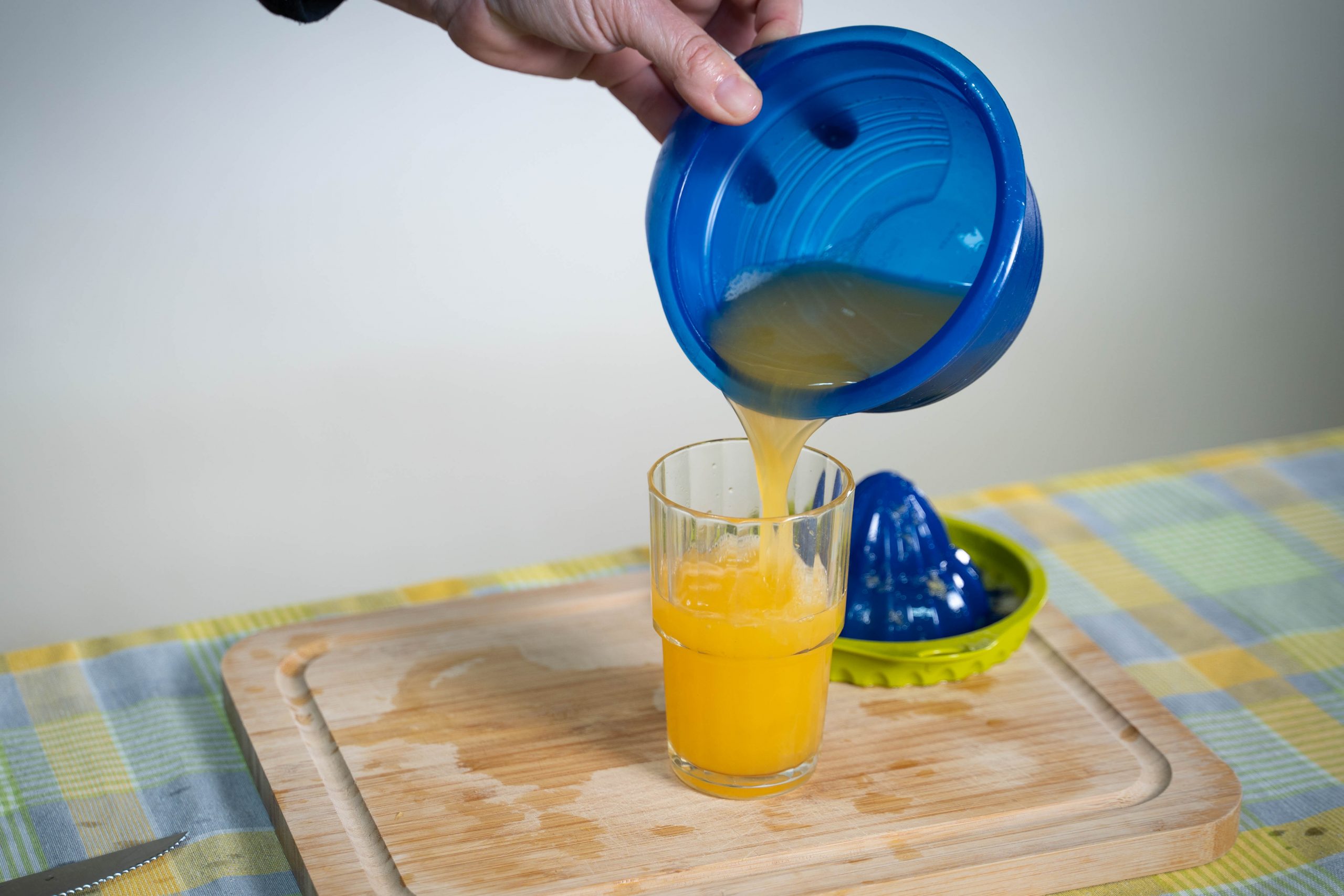 .שולחן עם קרש חיתוך ועליו כוס עם מיץ תפוזים. מעל הכוס יש יד ששופכת ממסחטה מיץ לכוס