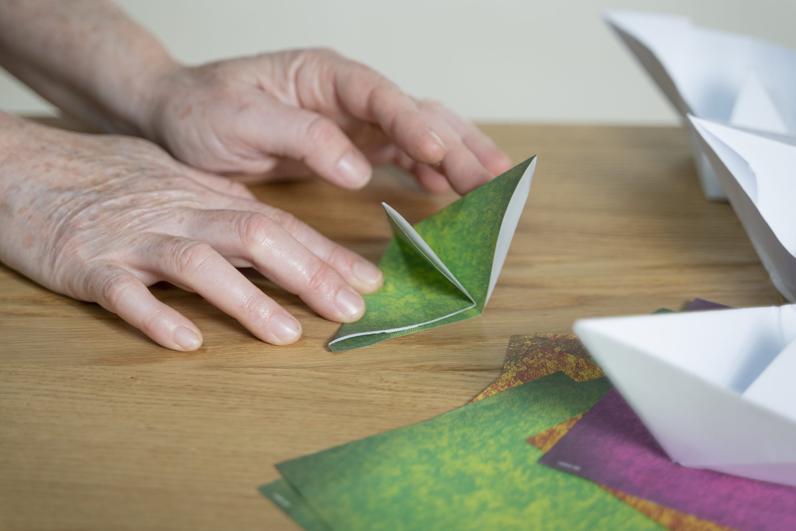 .זוג ידיים מקפלות נייר אוריגמי לצורה של שני משולשים. על השולחן ברקע ניירות אוריגמי צבעוניים ולבנים נוספים