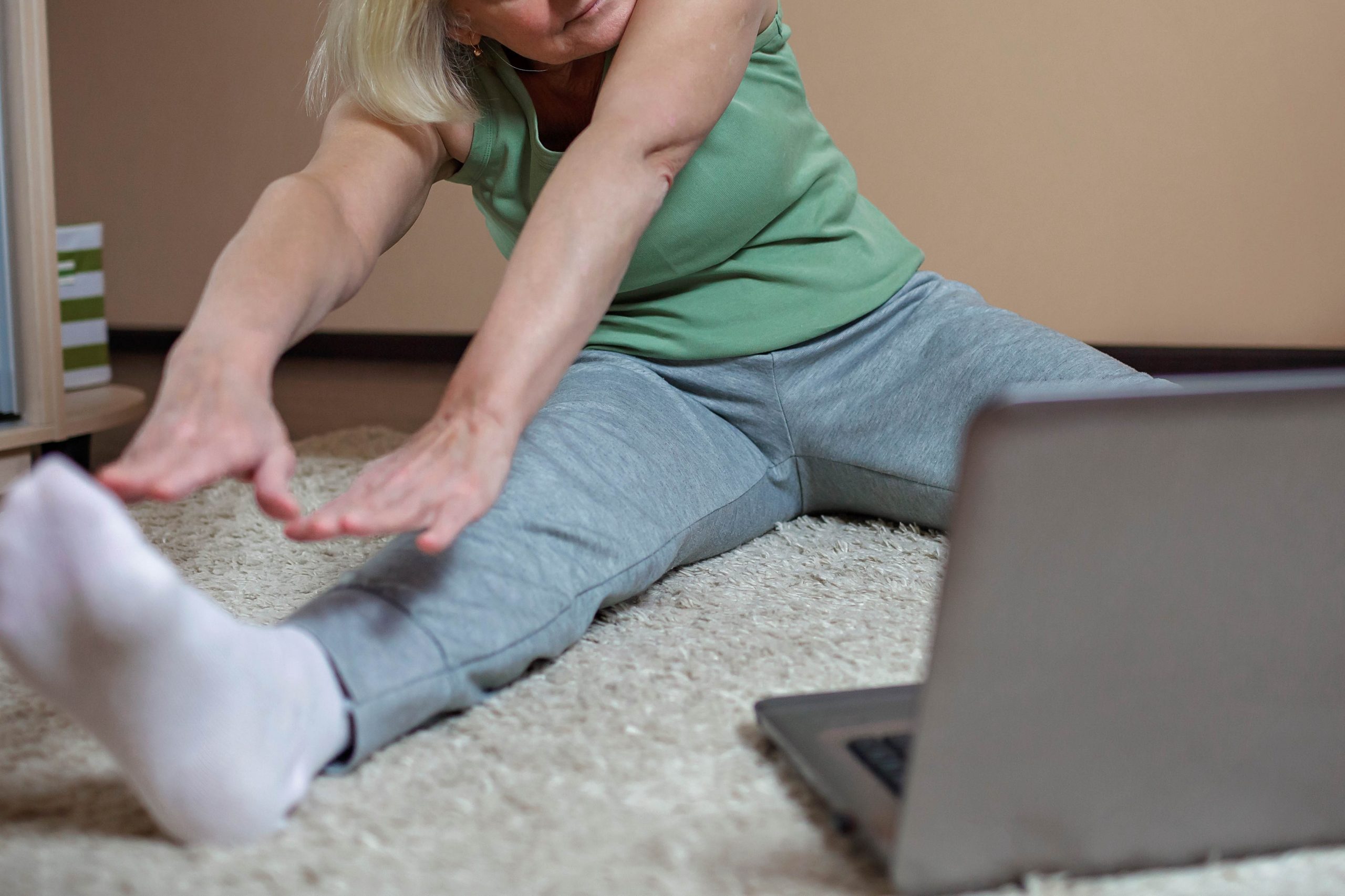 .אישה יושבת על שטיח ומחקה פעילות גופנית שיש במחשב הנייד שמולה. האישה יושבת בפיסוק ומותחת את שתי ידייה לכיוון אחת הרגליים