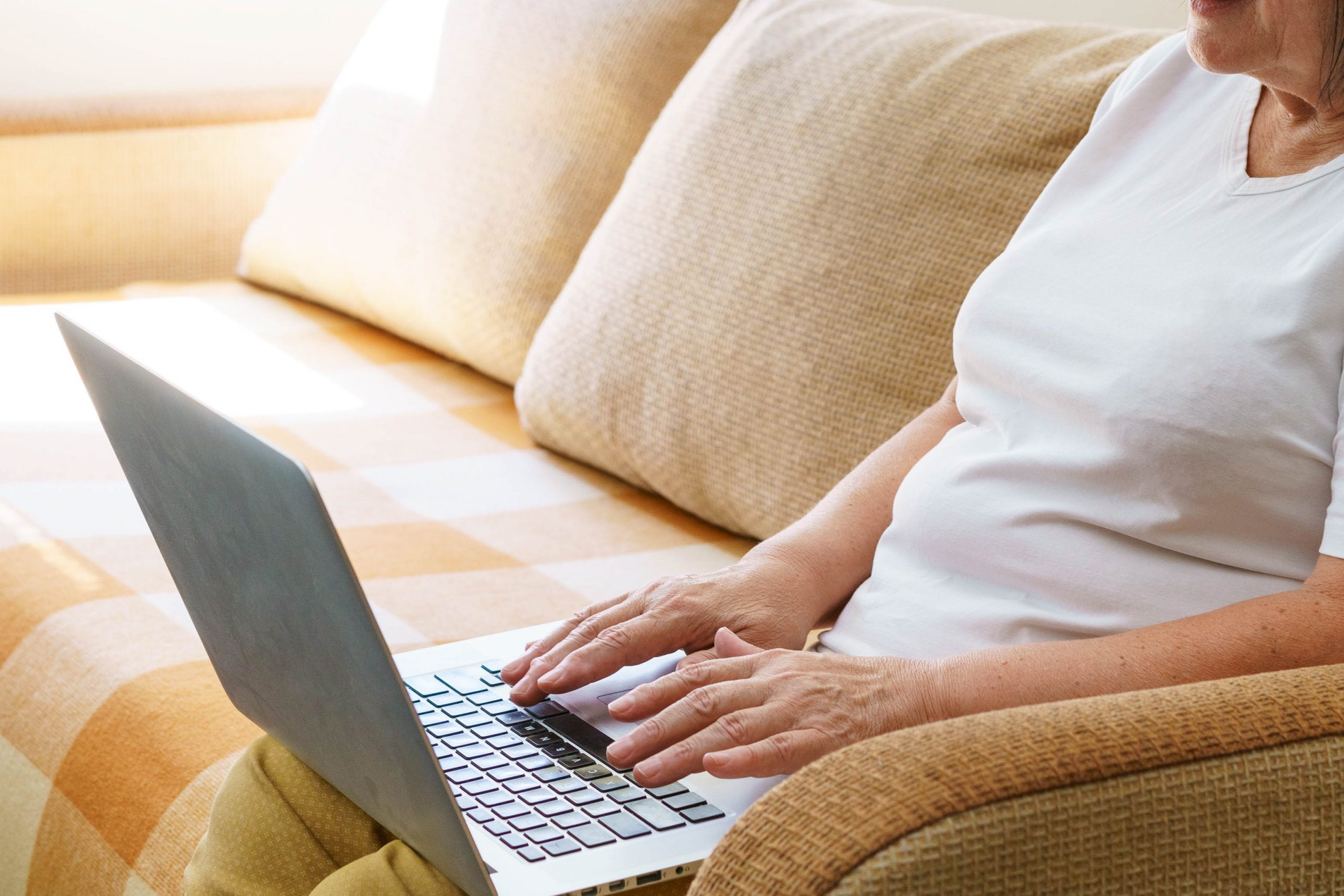 .אישה מבוגרת יושבת על ספה ומחזיקה על ברכיה מחשב נייד. ידיה מקלידות על גבי המחשב