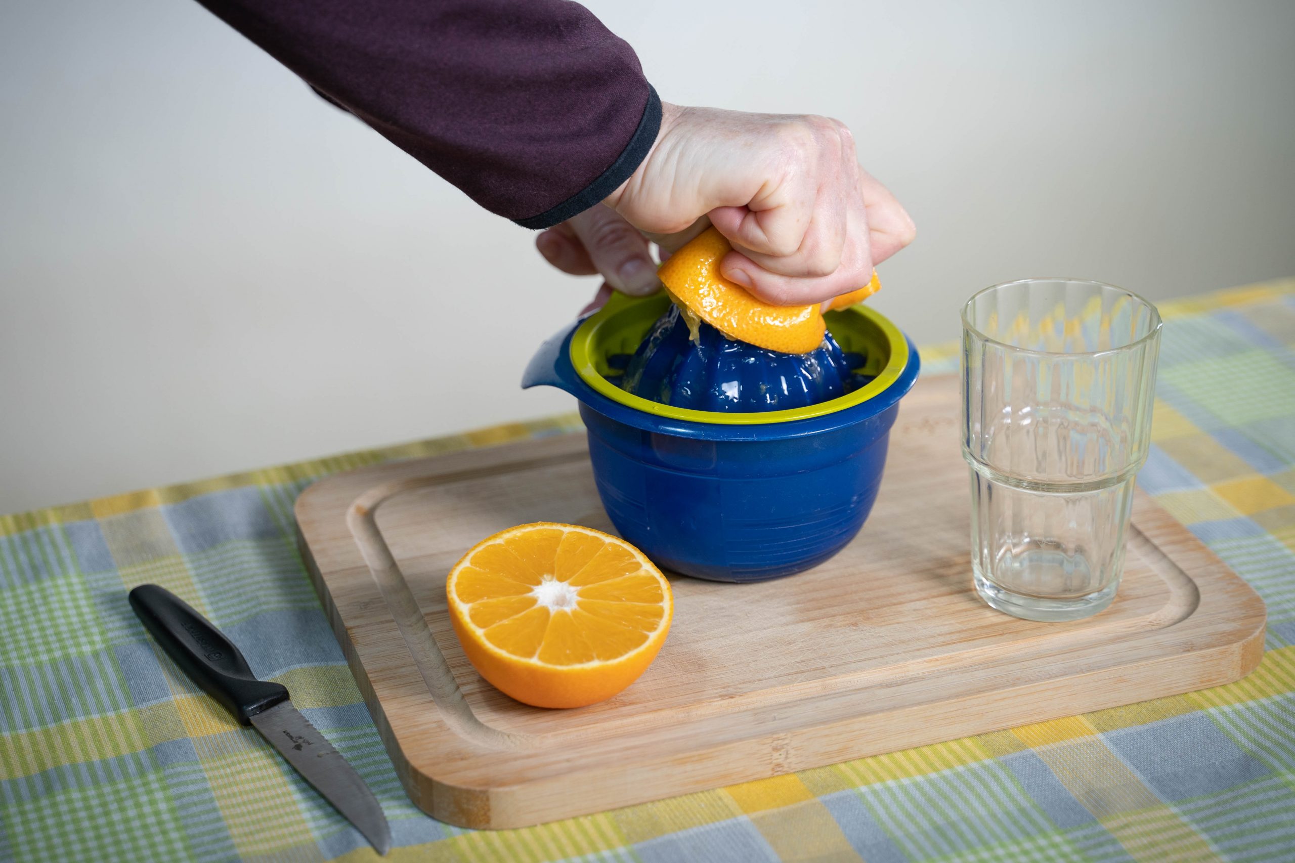 .יד סוחטת חצי תפוז במסחטה שנמצאת על קרש חיתוך על שולחן. לצד המסחטה, חצי תפוז נוסף, כוס זכוכית ריקה וסכין