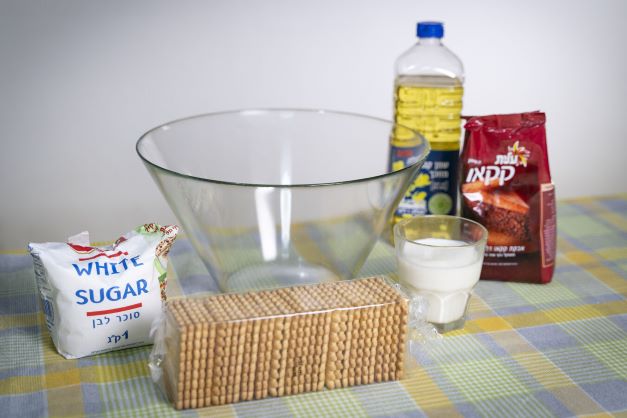 .שולחן ועליו מצרכים להכנת כדורי שוקולד - קערת זכוכית, סוכר, ביסקוויטים, כוס חלב, קקאו ושמן